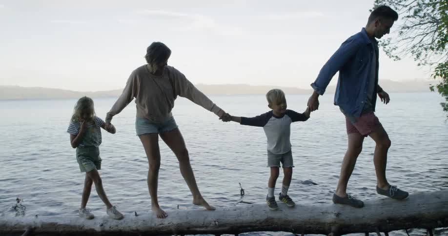 A family hold hands walking over a log on the water
