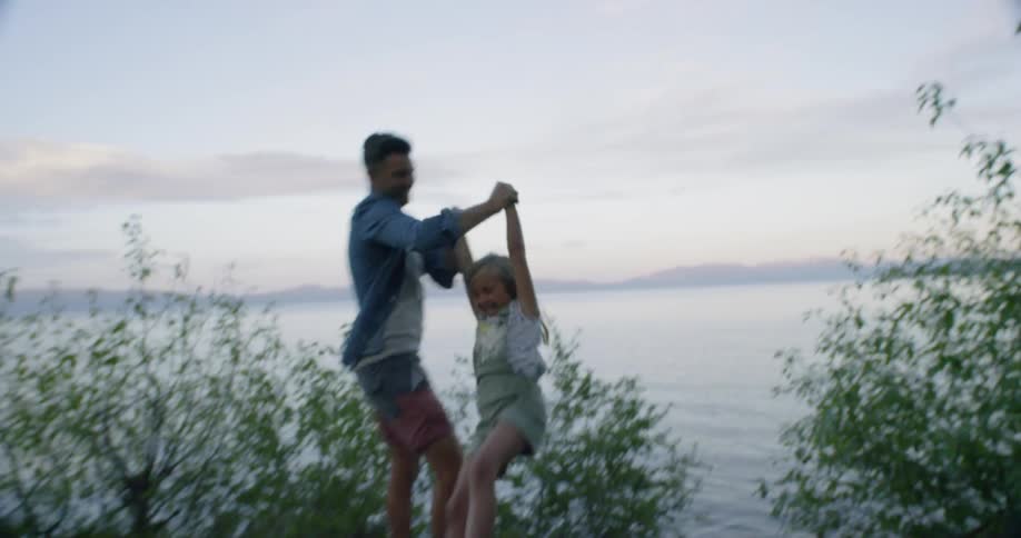 A father swings his daughter next to a lake at sunset