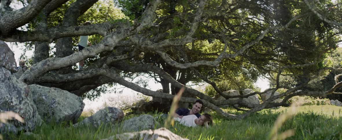 A man and woman lie in the grass in a natural setting as their children play near them