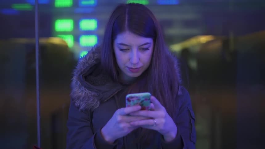 A young woman sits in front of a colorful display of lights while using her smartphone