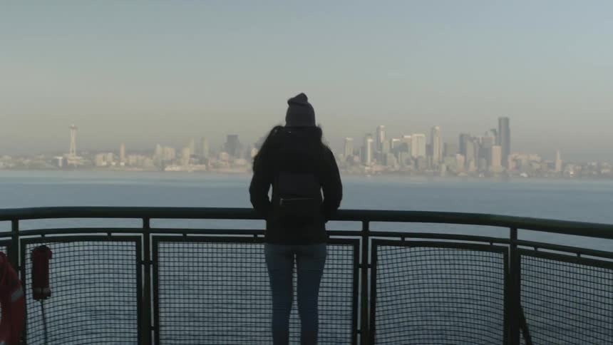 A woman is standing on the bow of a boat as it approaches a city in daylight
