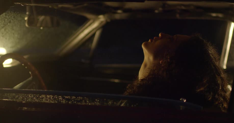 A woman leaning back in a car on a rainy night is sitting up