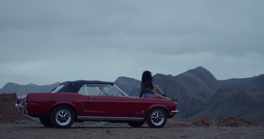 Woman sits on the hood of a red convertible in the country on a cloudy day