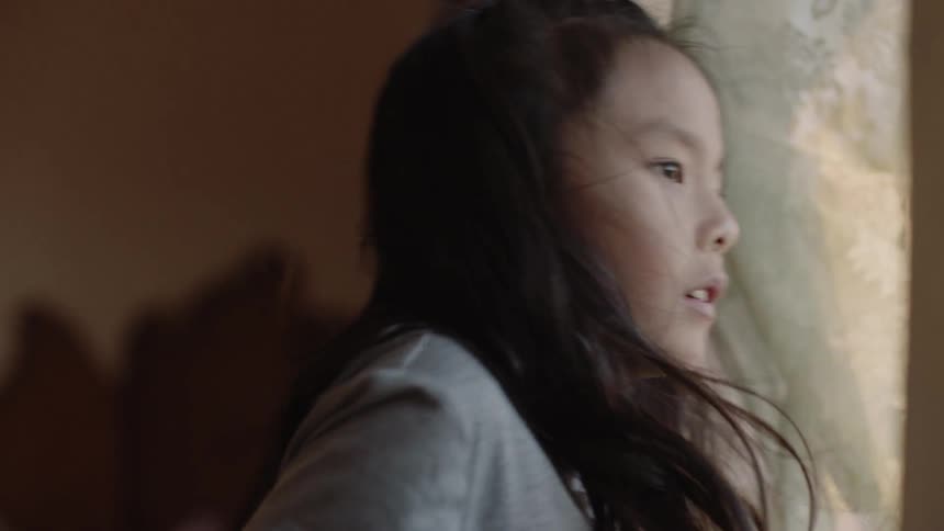 Child looks out a curtained window on a sunny day