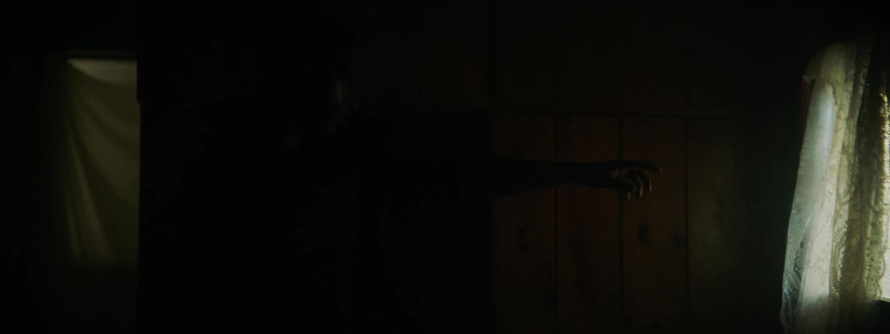 A man walks in a dark room and reaches towards a window