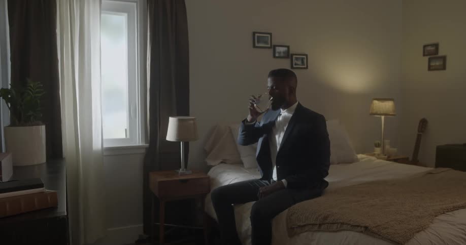 A man sitting on a bed buttons his collar and sips water