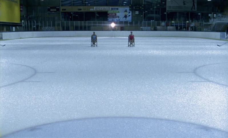 Two women sit in chairs on ice doing a choreographed routine as two hockey players skate by