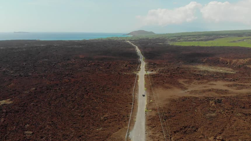 A vehicle travels on a road through barren land leading to the ocean