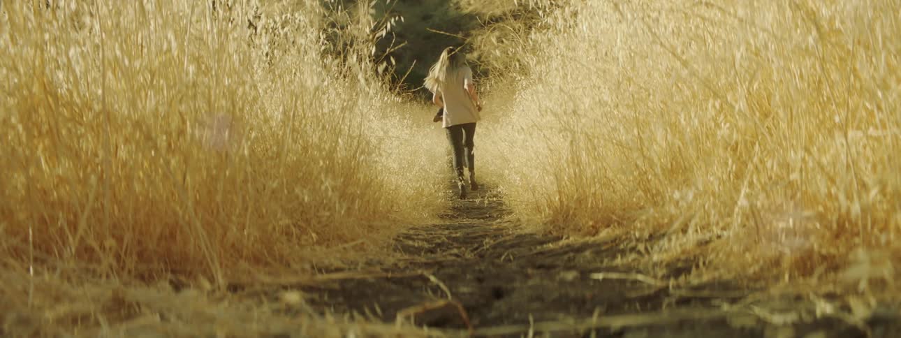 A man and woman run through a field of tall grass on a path