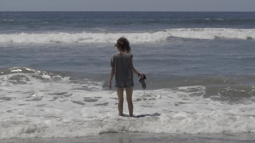 A young girl in jean shorts stands alone on the beach as the waves land at her feet