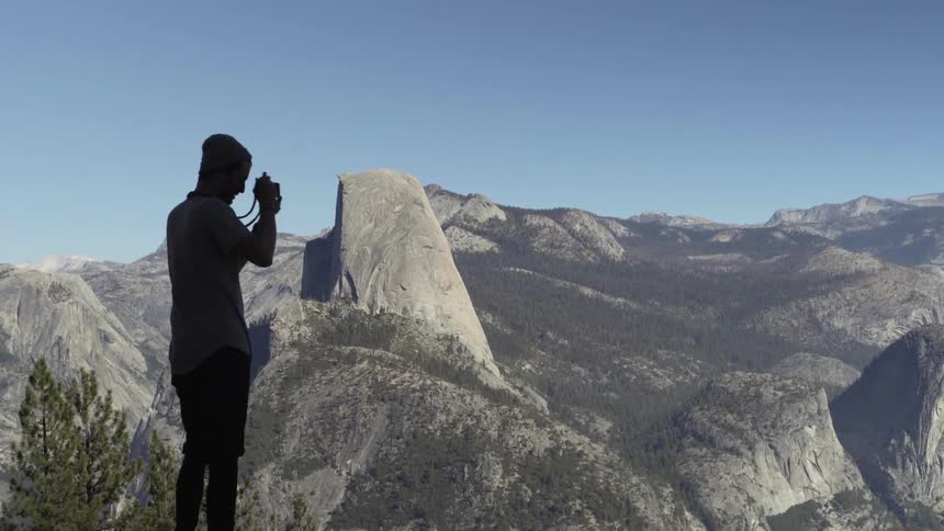 A man stand on a ledge and takes photographs of the landscape below