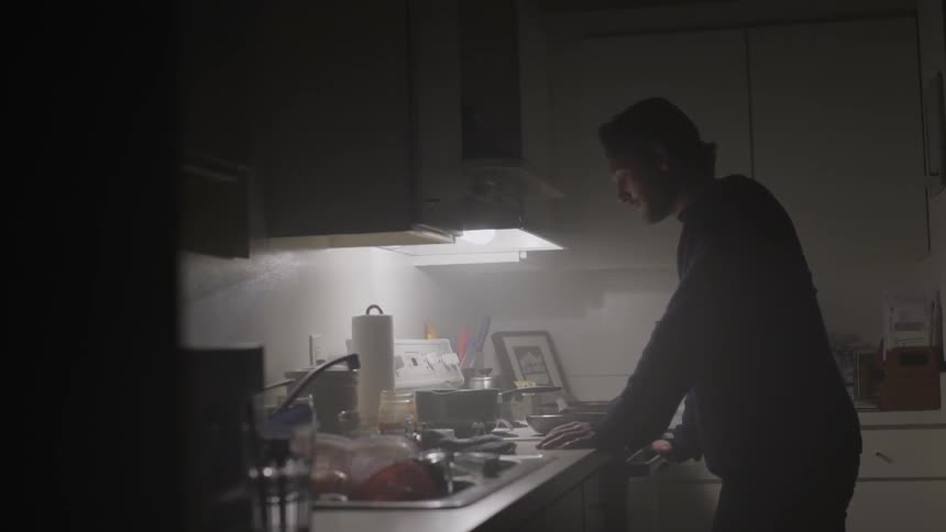 A man stares a stove top in a dimly lit kitchen