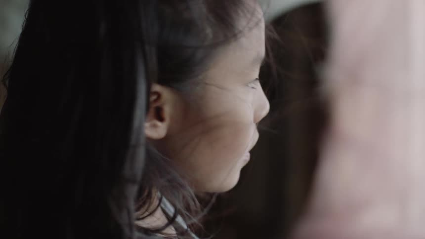 A little girl is holding back a curtain as she looks out a window on a windy day