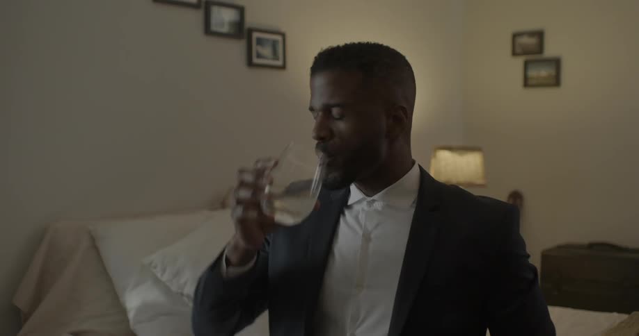 A man in a suit drinks wine while sitting on a bed