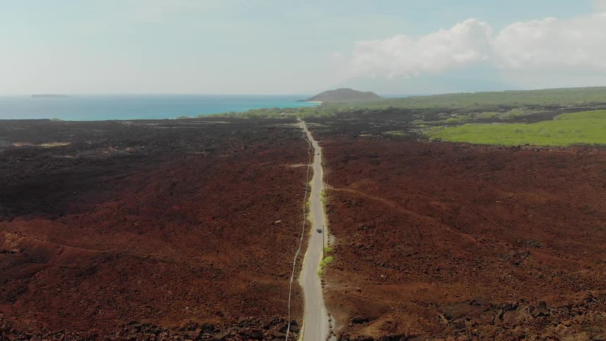 A car travels on an empty road through a rocky landscape towards the ocean