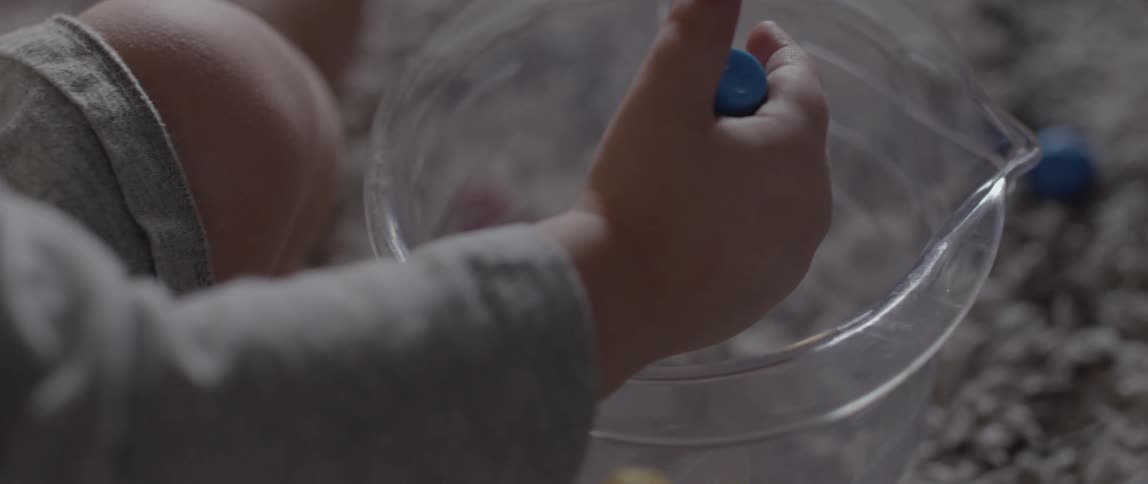 A toddler is dropping toy pegs into a mixing bowl in daylight