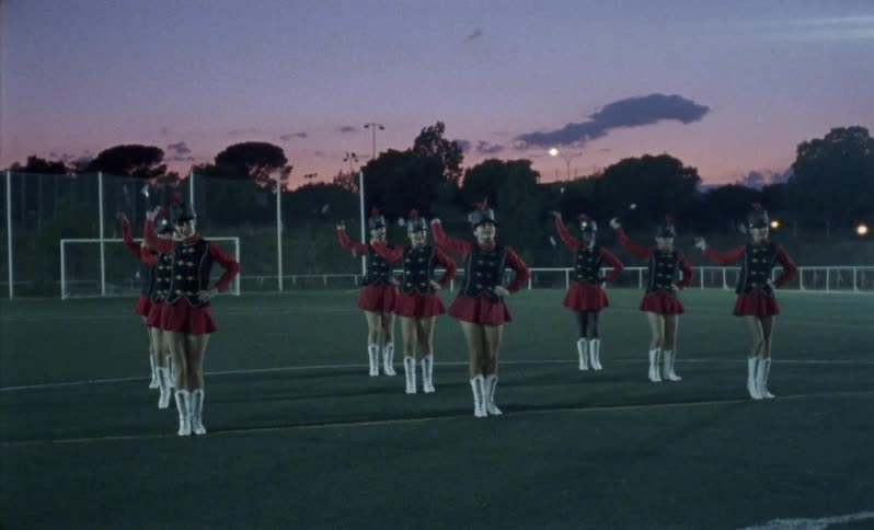 Girls in a baton corps are performing a routine on a football field at dusk
