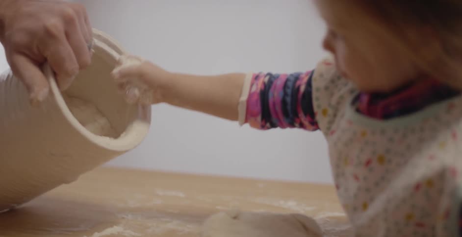 A little girl is sprinkling flour onto a ball of dough in a kitchen in daylight