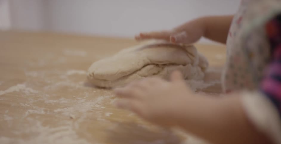 Child kneads flour covered dough on a wooden table