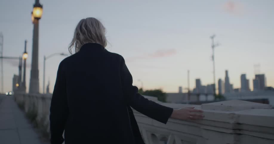 A woman is dragging her hand along a railing as she walks on a bridge at dusk