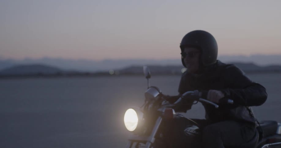 A man leans forward as he ride a motorcycle in the desert at sunset