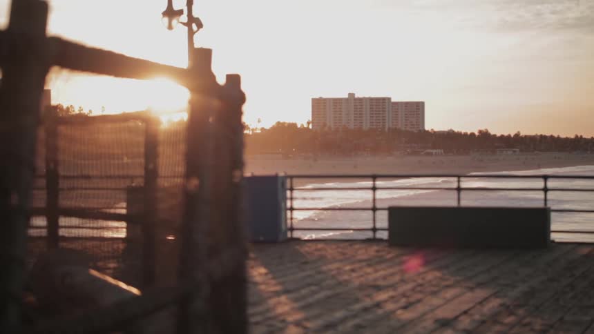 View from a pier of waves rolling to shore and the sun setting behind a hill and some buildings