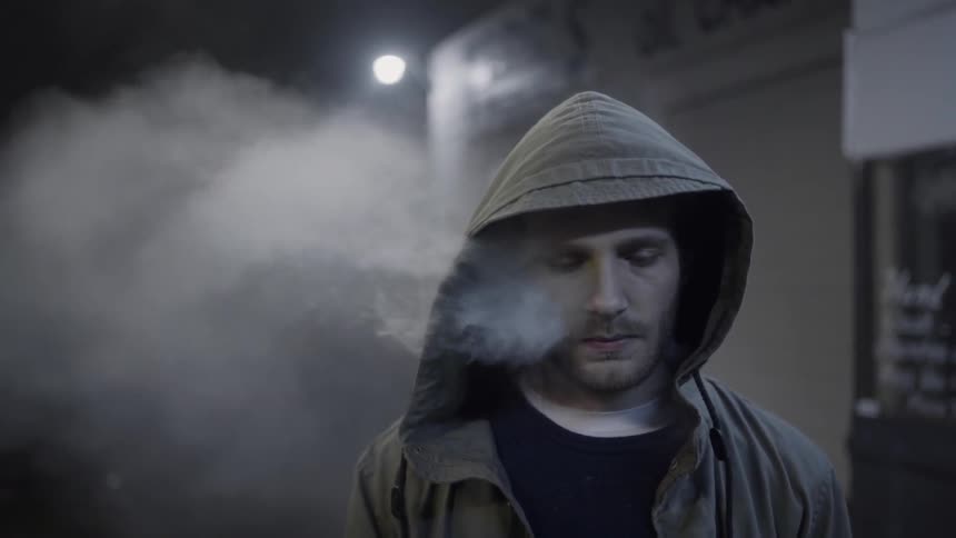 A man wearing a hoodie stands on a sidewalk at night with his eyes closed as steam billows from within the hood of his jacket