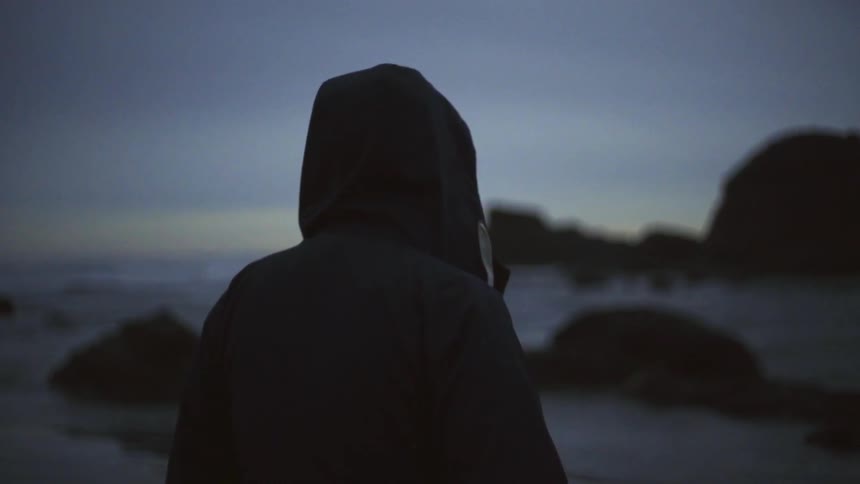 A person in a coat stands near the water at dusk