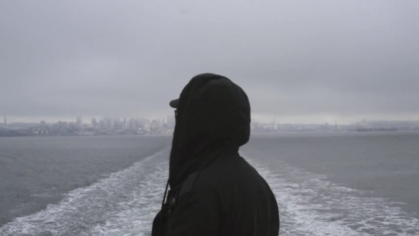 A man stands at the rear of a boat while viewing a skyline as the boats goes out to sea