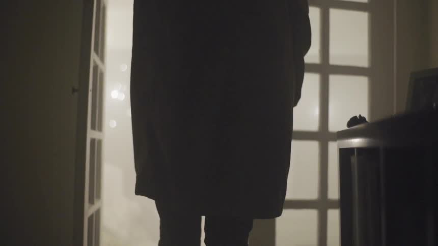 A man wearing a trench coat opens a french door and begins walking out of the room