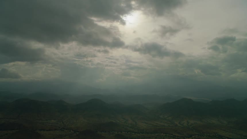 The sun is casting rays through clouds as they hang over a mountain range