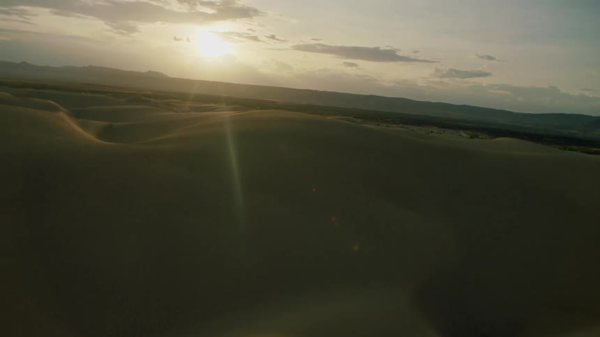 The sun is shining on the horizon of a desert landscape beyond sand dunes