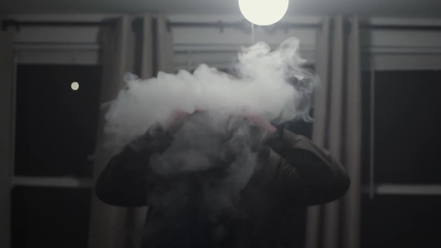 A man wearing a hoodie stands up inside a room while exhaling a large amount of smoke