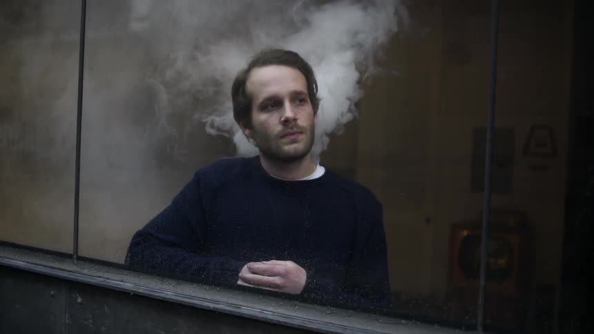 A man standing in front of a window as smoke starts to billow from his collar