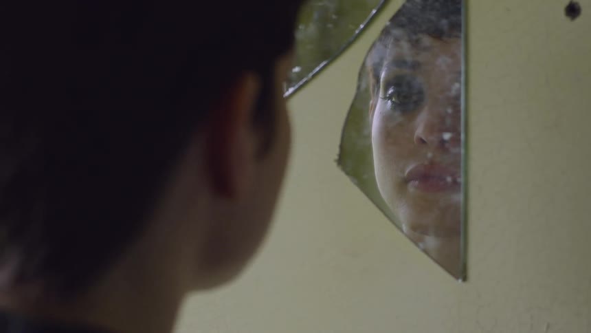 Woman looks at her face in a broken piece of mirror and wipes her eyes