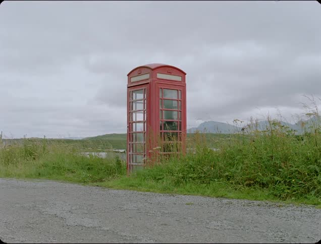 A red telephone booth on the side of the road surrounded by grass