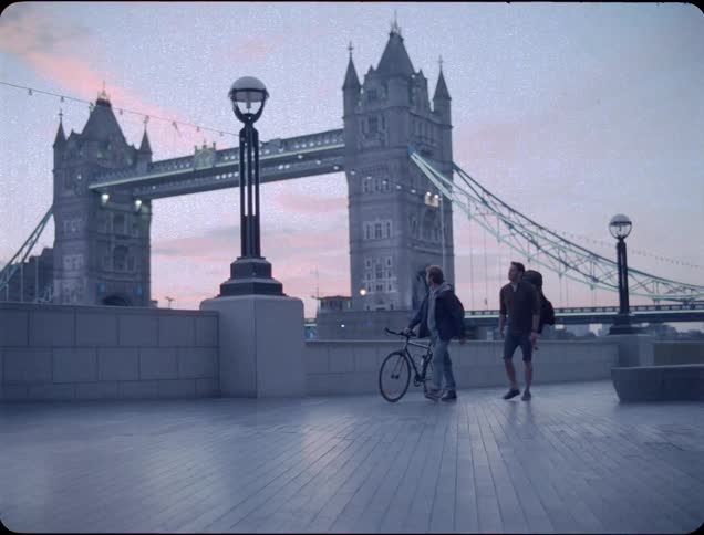 A man pushing a bike hugs a friend as they walk near a bridge at dawn