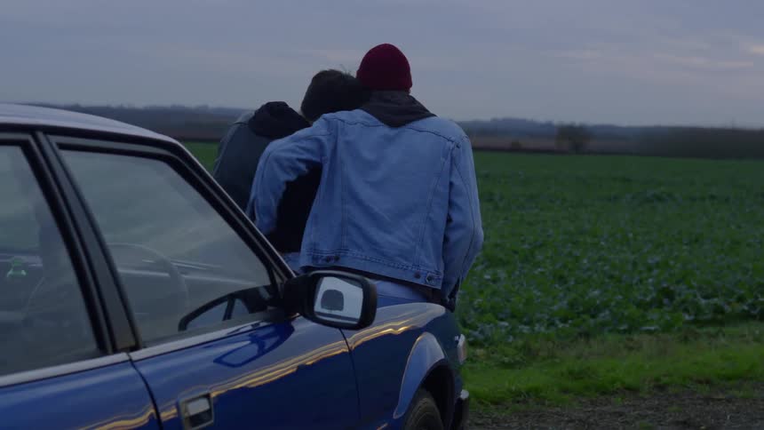 Man and woman sit on a car hood and the man puts his arm around the woman