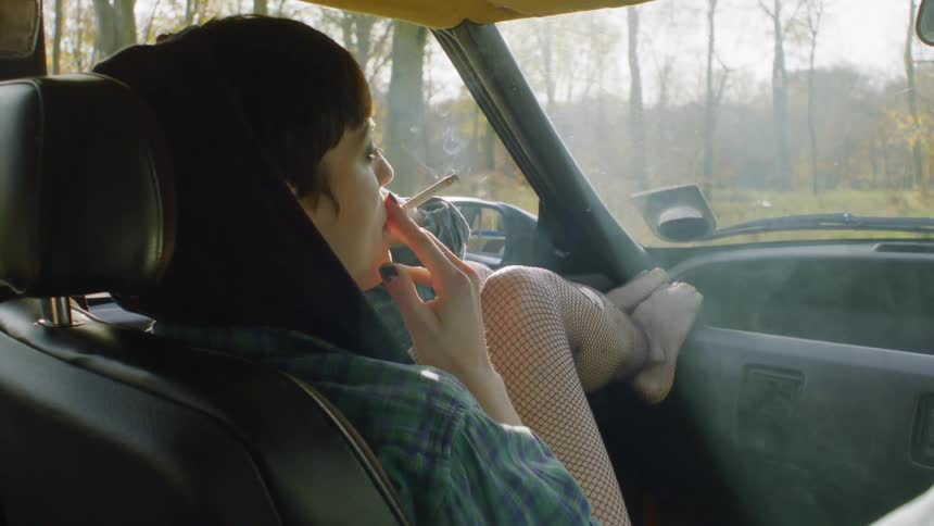 Woman smokes a cigarette as she sits in a car in the forest