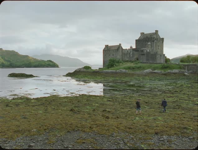 People walking in a marshy area near an old building on a cloudy day