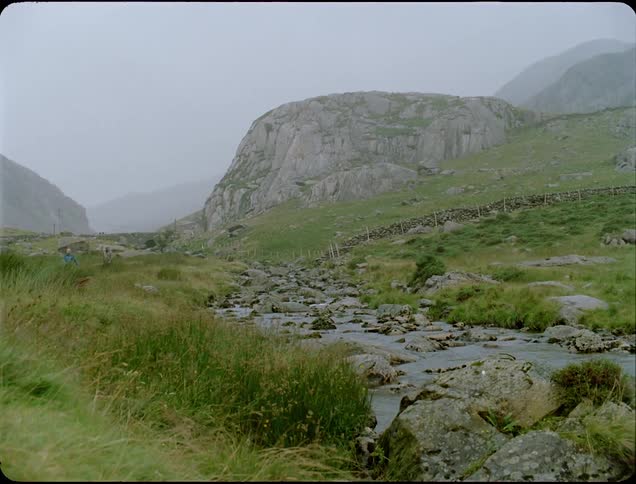 Two people walk on grassy land near a rocky stream