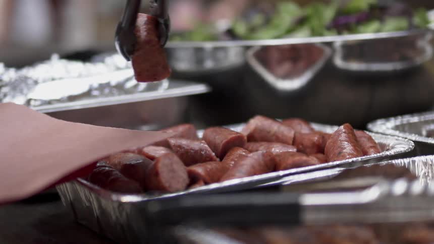 A person uses tongs to lift up sausage onto a plate