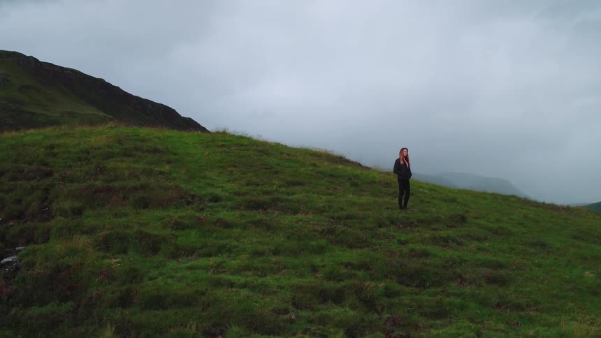 A woman walking down a hillside on a cloudy day is stopping to admire the view