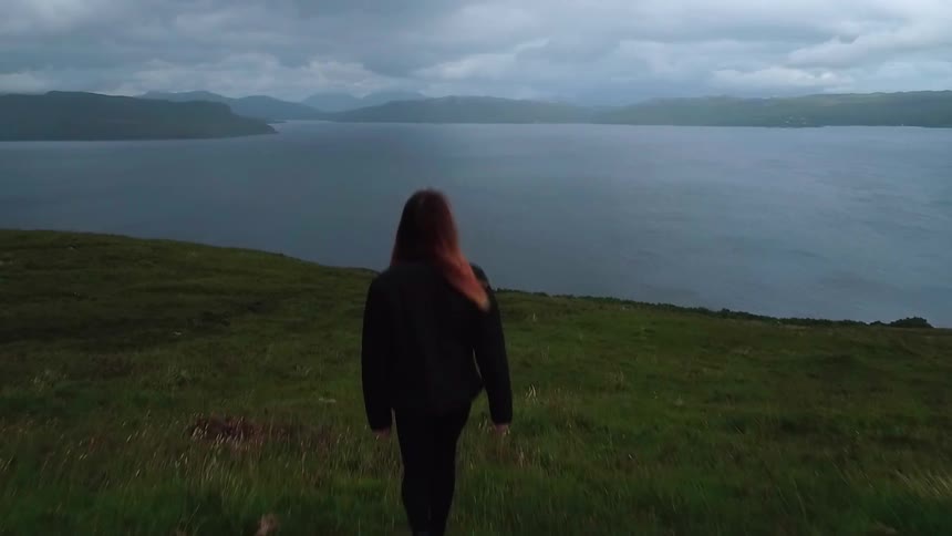 A woman walks through grass towards a large body of water under a cloudy sky
