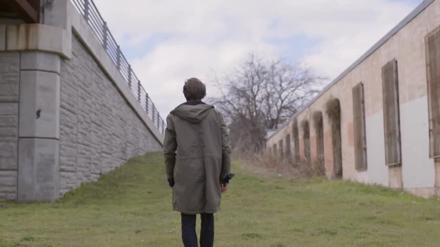 A man wearing a stadium length coat standing and turning around in between a highway ramp and an old brick building