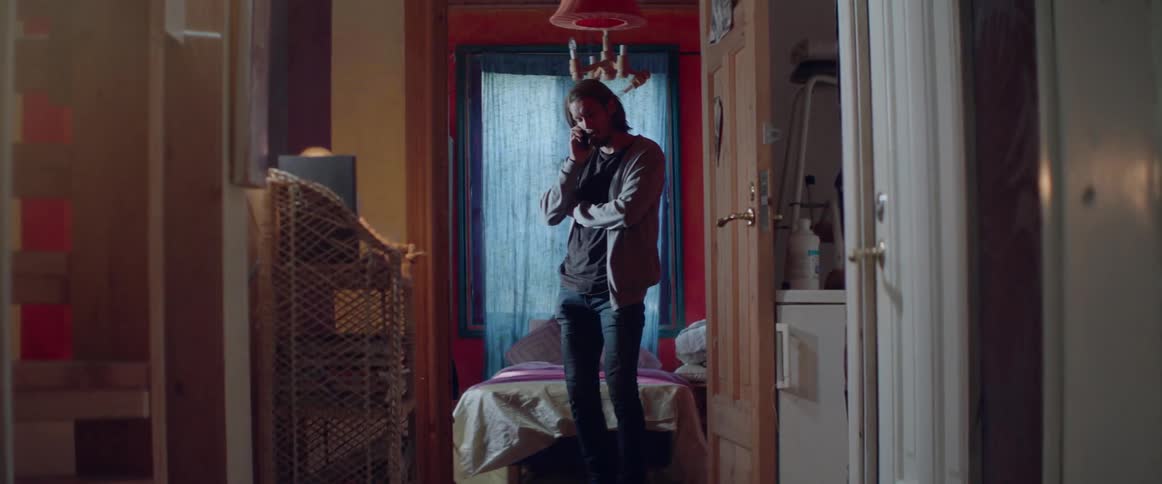 A man stands near a wooden doorway having a mobile phone conversation inside.
