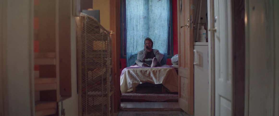 A serious young man sits cross legged on bed during phone conversation.