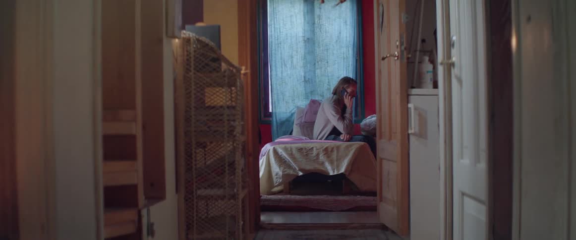 A view down a hallway shows a man sitting sideways on his bed.