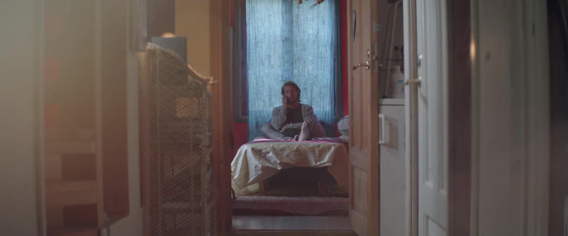A serious male character sits cross legged on his bed having phone call.