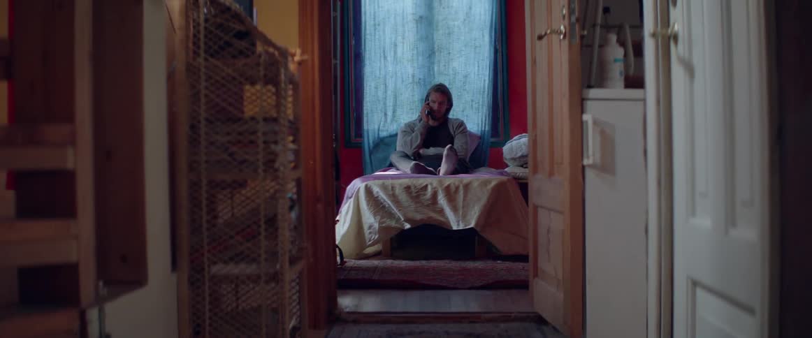 Wide shot of a male character holding a mobile phone while sitting indoors.